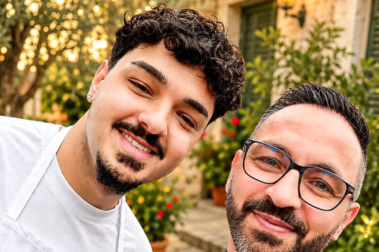 Zwei lächelnde Männer mit Schürzen posieren für ein Selfie vor einem Innenhof mit Lichterketten.