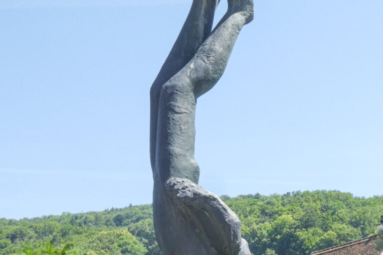Eine Bronzestatue zeigt einen Menschen kopfüber mit erhobenen Beinen, umgeben von blühenden Sträuchern vor Waldhügeln.