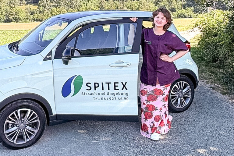 Eine Frau lehnt lächelnd an einem weissen Spitex-Auto vor einer Hügellandschaft.