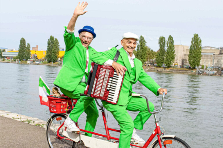 Zwei Männer in grünen Anzügen fahren auf einem roten Tandem am Flussufer, einer winkt, der andere spielt Akkordeon.