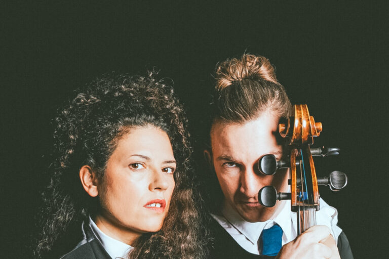 Eine Frau mit lockigem Haar hält ein Keyboard, dahinter ein Mann mit Dutt hält ein Cello.