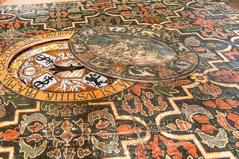 Bemalte Tischplatte mit ornamentalem Rankenmuster, lateinischer Inschrift und rundem Landschaftsmedaillon mit Wappen.
