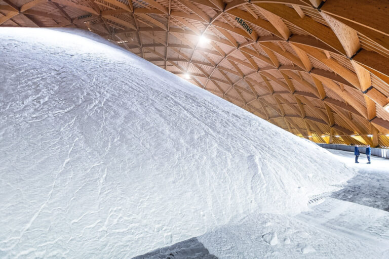 Ein riesiger Salzhaufen füllt eine hölzerne Lagerhalle mit gewölbtem Dach, zwei kleine Personen am Rand.
