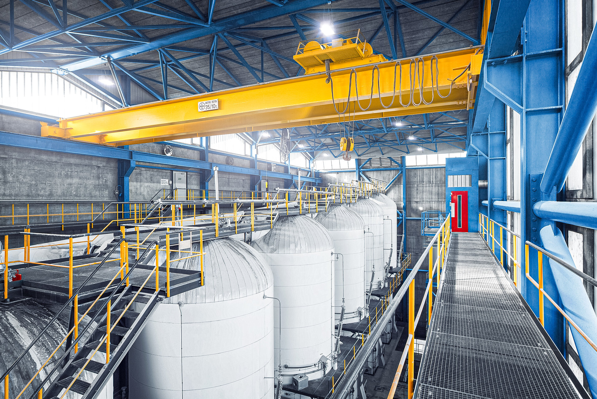 Industriehalle mit Reihe weisser Rundtanks, darüber ein gelber Kran an blauer Stahlkonstruktion.