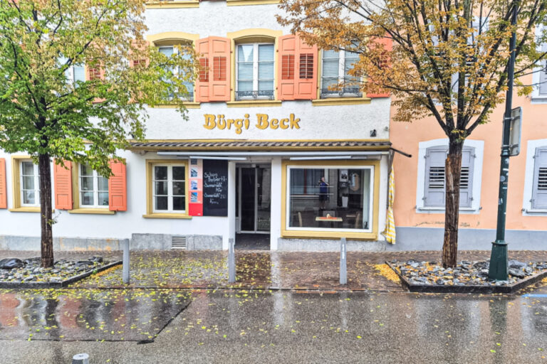 Fassade einer Bäckerei mit Schriftzug „Bürgi Beck“ an einer regennassen Strasse mit herbstlichen Bäumen.