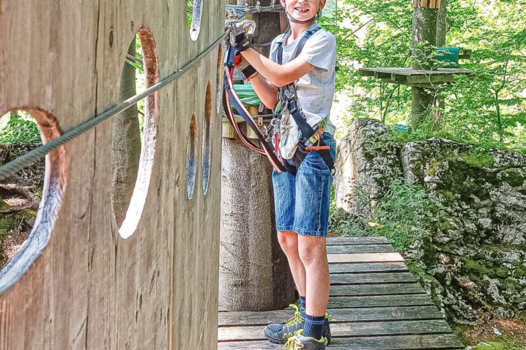 Ein Junge mit Helm und Klettergurt steht gesichert auf einer Plattform im Hochseilgarten.