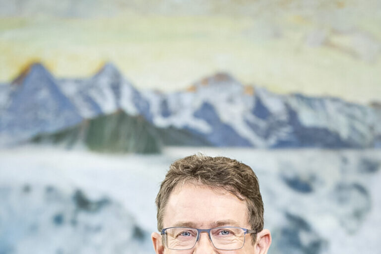 Ein lächelnder Mann mit Brille und blauem Anzug samt Krawatte steht vor einem Gemälde mit Berglandschaft.