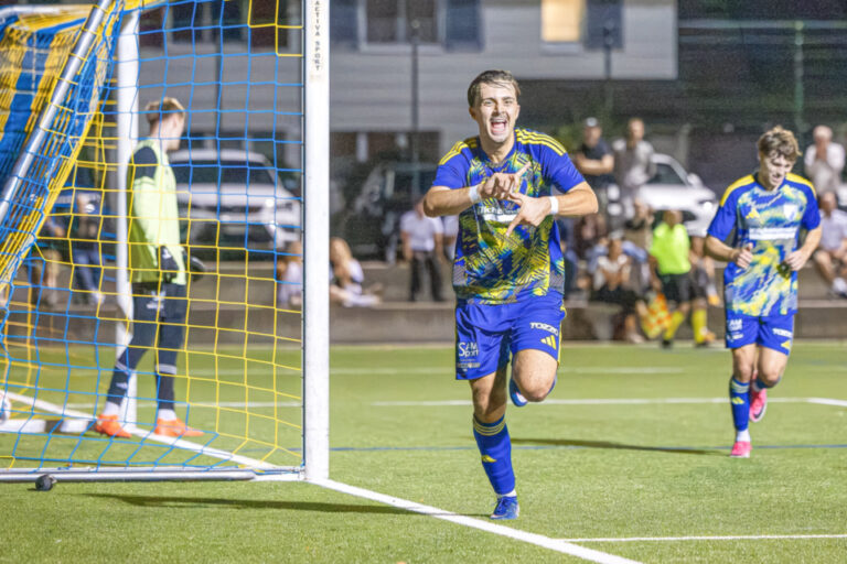Ein jubelnder Fussballspieler in blau-gelbem Trikot rennt lachend über den Platz, im Hintergrund der Torhüter und Zuschauer.