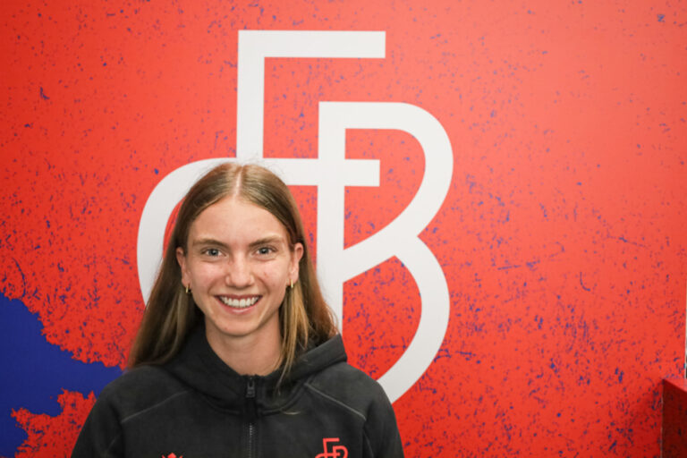 Eine lächelnde junge Frau mit langen Haaren steht vor einer roten Wand mit grossem Logo.