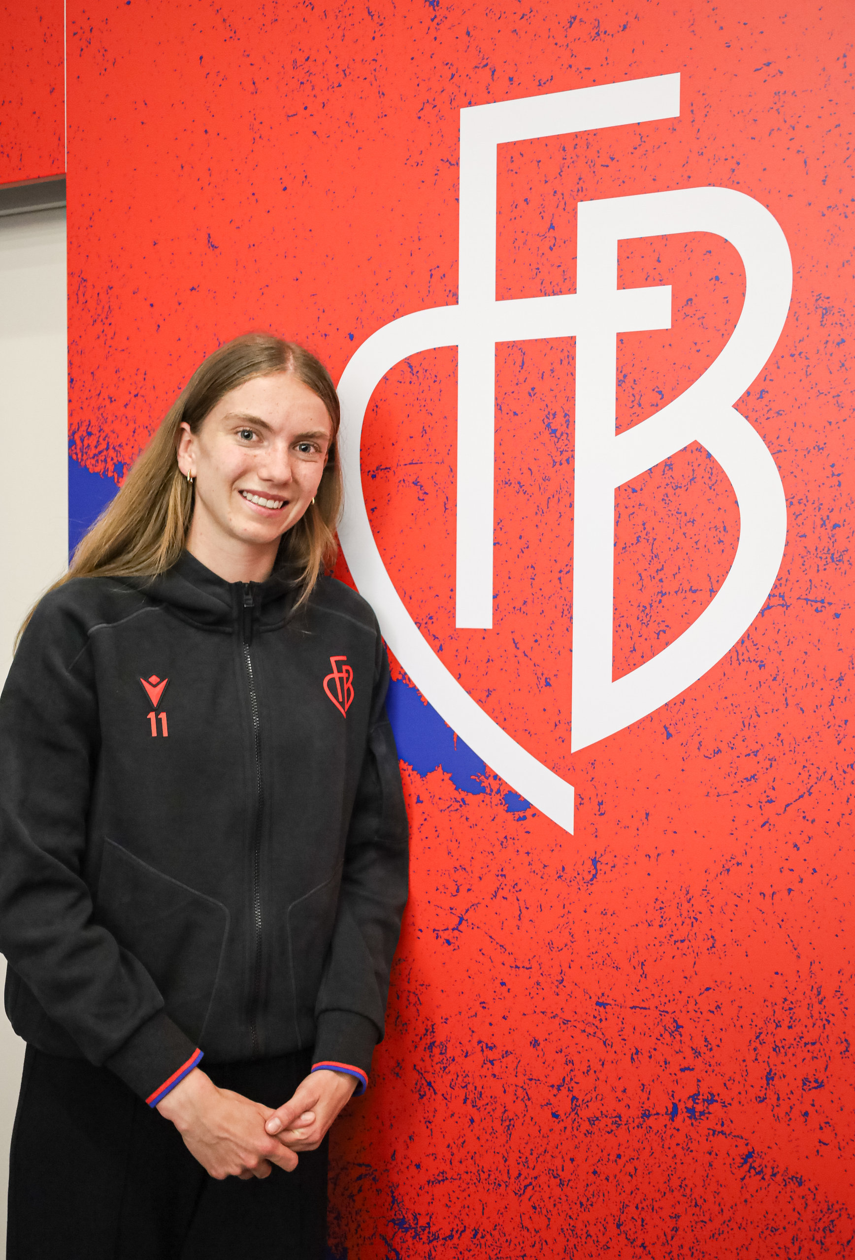 Eine lächelnde junge Frau in schwarzer Trainingsjacke steht vor einer roten Wand mit einem grossen weissen Vereinslogo.