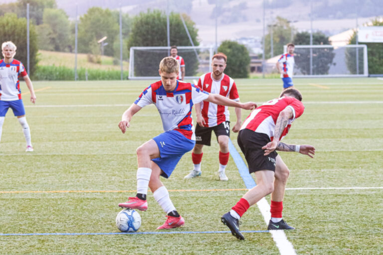 Fussballspieler kämpft am Ball gegen einen Gegenspieler, weitere Spieler im Hintergrund auf dem Rasen.