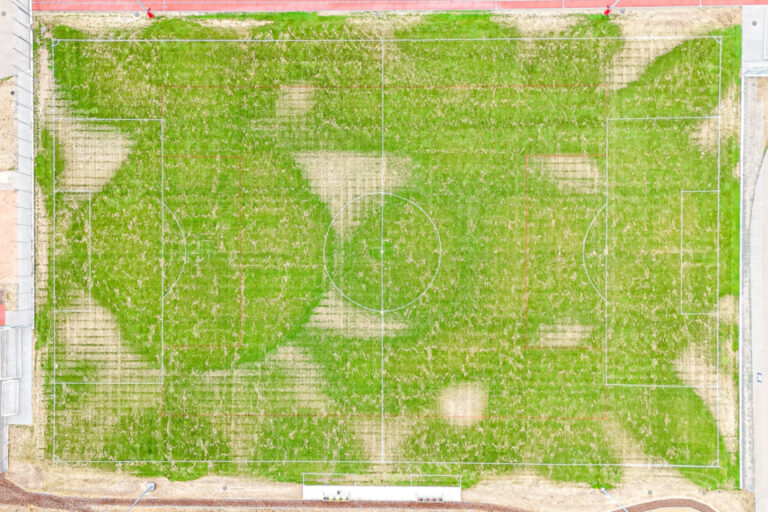 Luftaufnahme eines Fussballfeldes mit ungleichmässig gewachsenem, geflecktem Rasen zwischen roten Gebäudedächern.