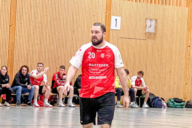 Ein Handballspieler im roten Trikot mit Nummer 20 geht über das Spielfeld, im Hintergrund sitzen Zuschauer.
