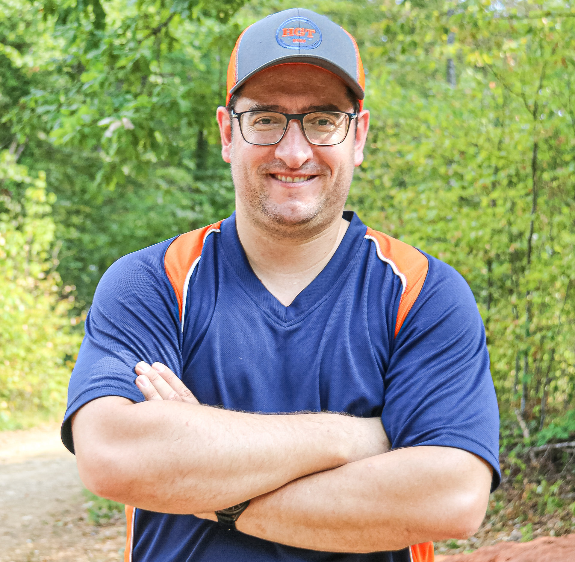 Ein lächelnder Mann mit Cap und Brille steht mit verschränkten Armen auf einem Waldweg.