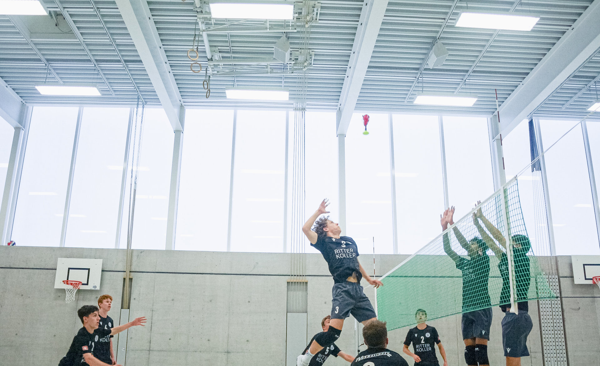 Volleyballspieler springt zum Angriffsschlag, gegnerische Spieler blocken am Netz in einer Sporthalle.