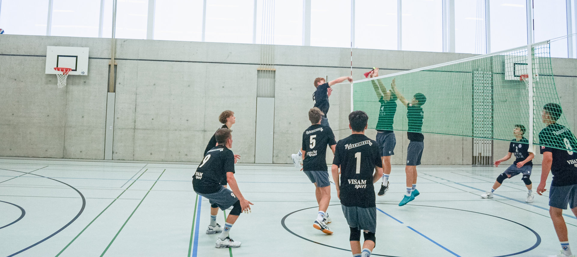 Jugendliche spielen Volleyball in einer Sporthalle, ein Spieler springt zum Schmetterschlag am Netz.