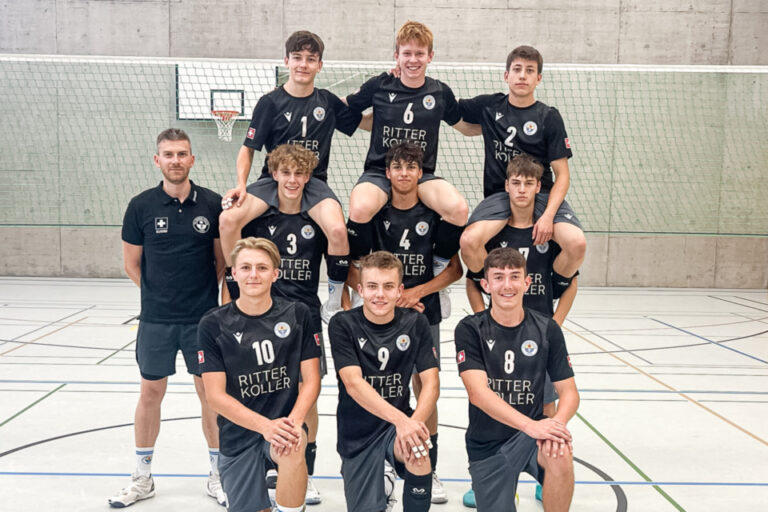 Eine Jugend-Volleyballmannschaft in schwarzen Trikots posiert in drei Reihen in einer Sporthalle.