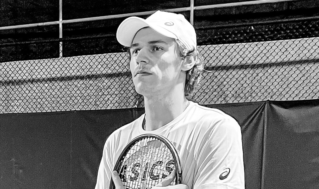 Ein junger Mann mit weisser Kappe hält einen Tennisschläger vor der Brust und blickt zur Seite.