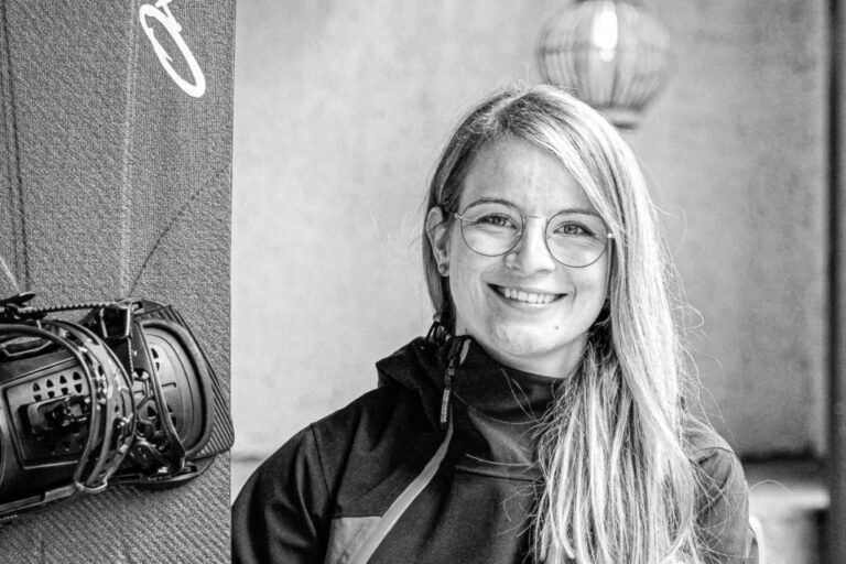 Lächelnde blonde Frau mit Brille sitzt auf einer Bank neben einem Snowboard mit Fernbedienung.