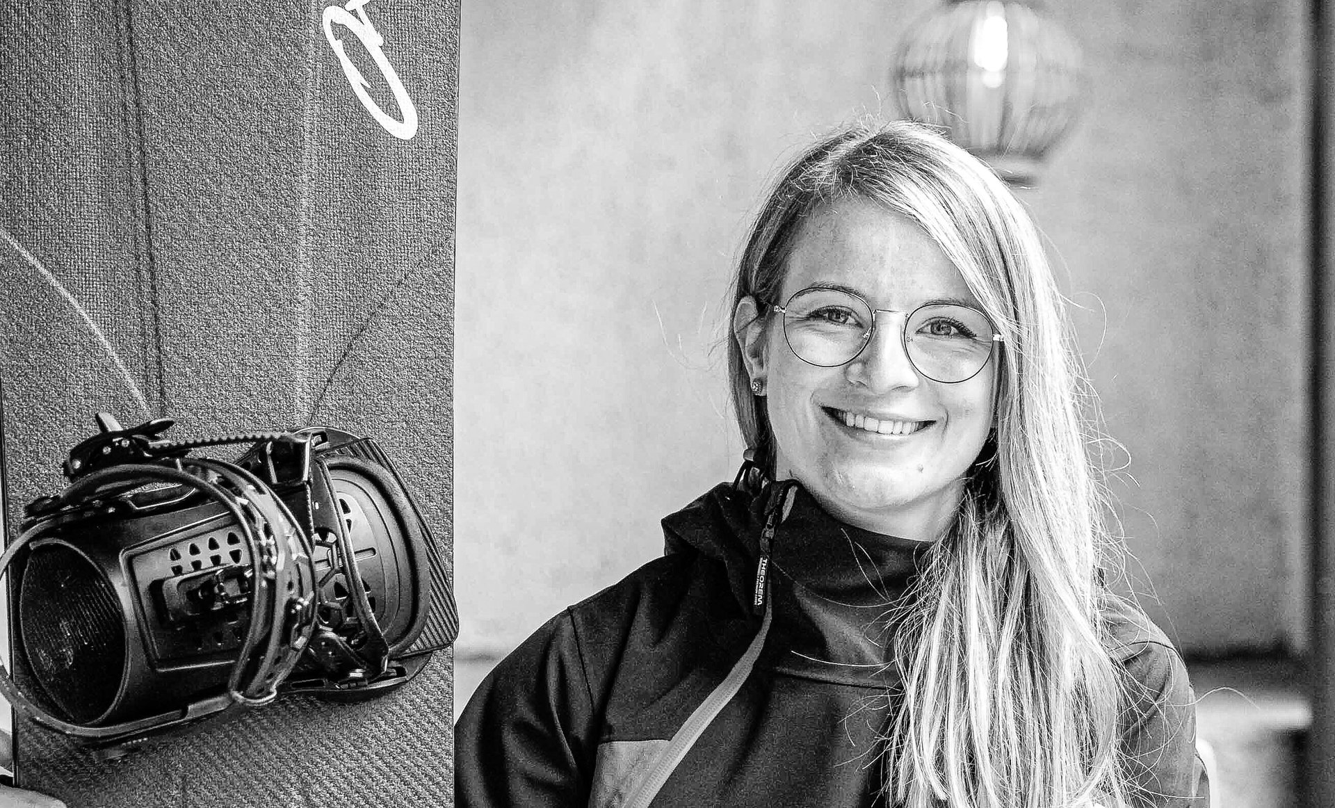 Lächelnde blonde Frau mit Brille sitzt auf einer Bank neben einem Snowboard mit Fernbedienung.