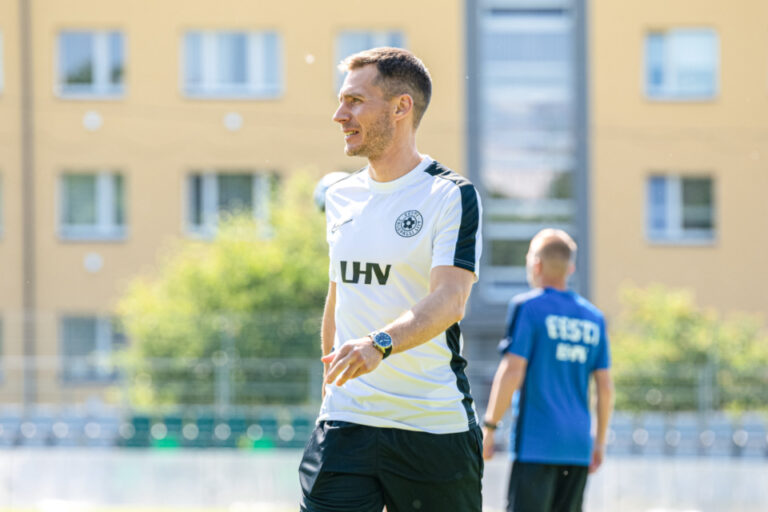 Ein Mann im Trainingsshirt lächelt und blickt zur Seite auf einem Sportplatz, im Hintergrund ein weiterer Mann.