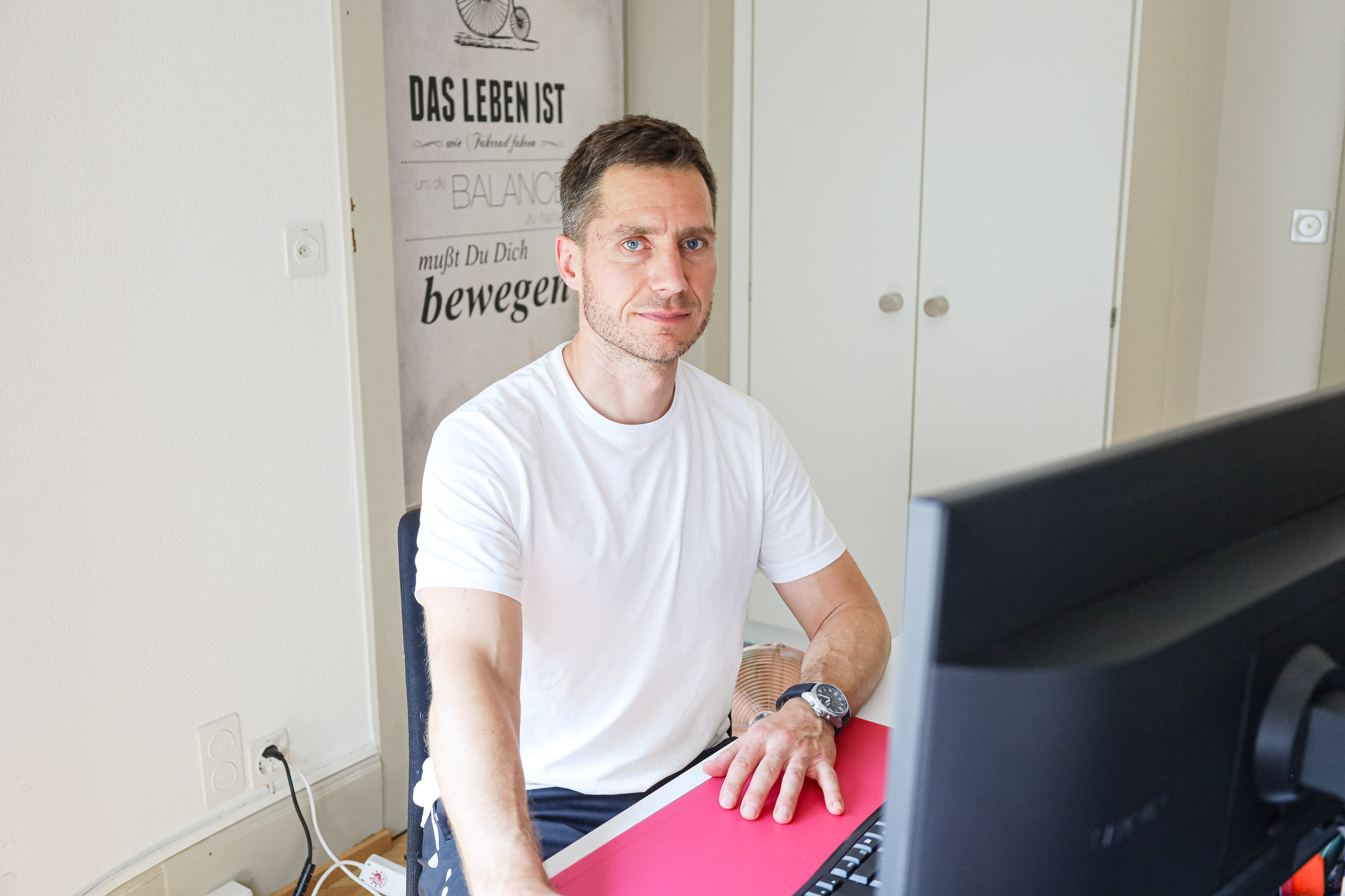 Ein Mann im weissen T-Shirt sitzt an einem Schreibtisch vor einem Computerbildschirm und blickt in die Kamera.