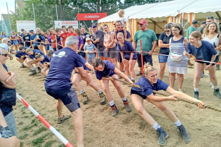Mehrere Personen in blauen Trikots ziehen beim Tauziehen an einem Seil auf sandigem Boden.