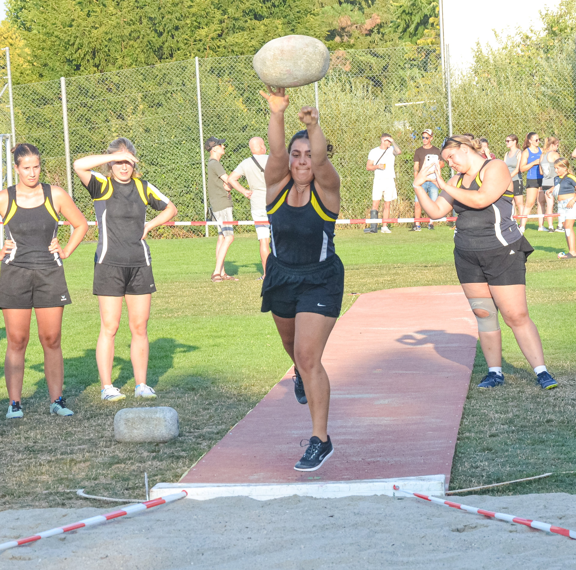 Eine Athletin holt zum Wurf mit einer schweren Kugel über dem Kopf aus, umringt von wartenden Sportlerinnen.