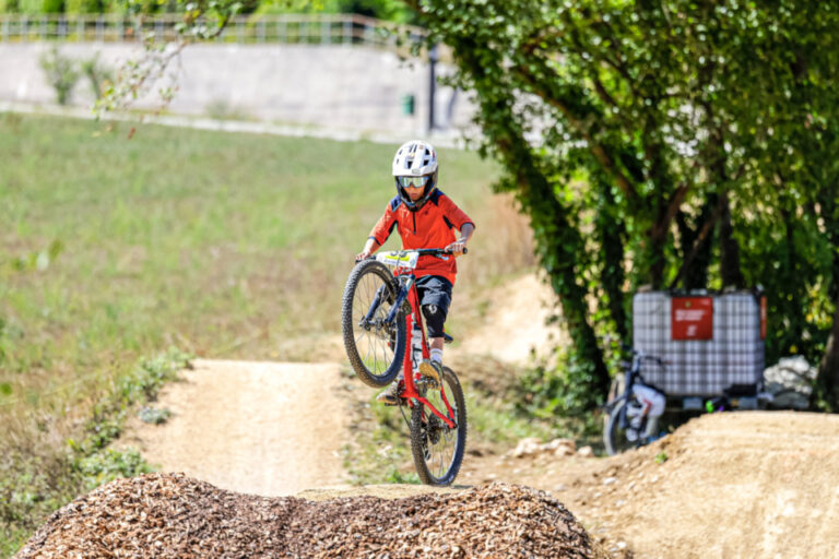 Ein Kind mit rotem Trikot und Helm springt mit dem Mountainbike über einen Erdhügel auf einer Bikepark-Strecke.