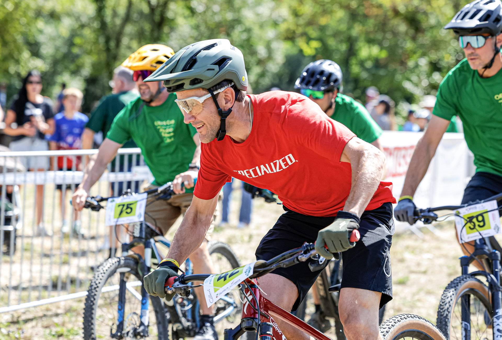 Mountainbiker mit Helmen und Startnummern fahren bei einem Rennen im Gelände, ein Mann im roten Trikot vorne.
