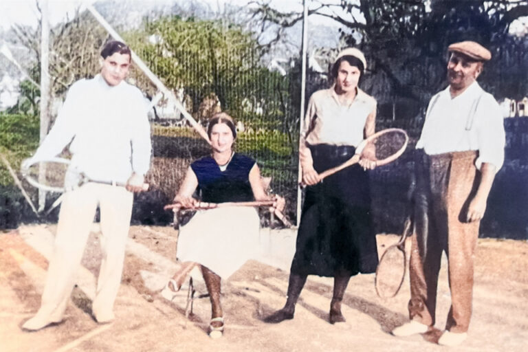 Vier Personen in altmodischer Kleidung stehen mit Tennisschlägern auf einem Sandplatz.