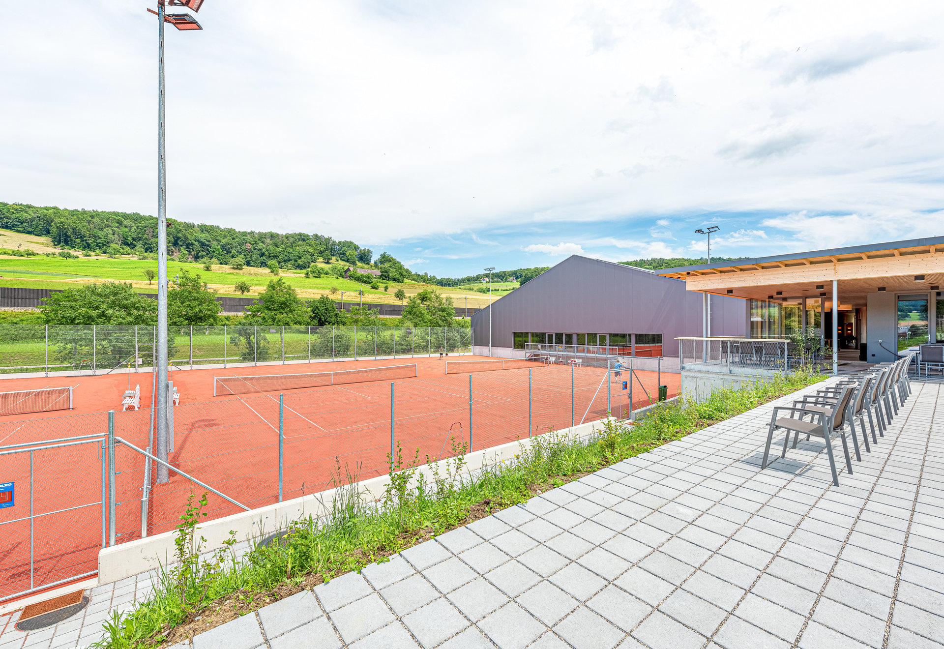 Tennisplatz mit Flutlicht neben einem modernen Vereinsgebäude und Terrasse mit Stühlen, umgeben von grünen Hügeln.