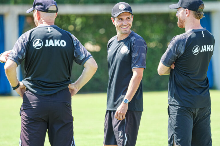 Drei Trainer in dunklen Trikots mit Cap stehen lachend auf einem Fussballplatz zusammen.