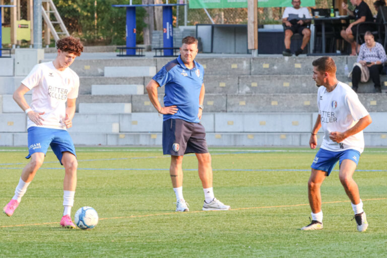 Fussballspieler und ein Trainer in Trainingskleidung auf dem Rasen, im Hintergrund Zuschauer auf einer Tribüne.