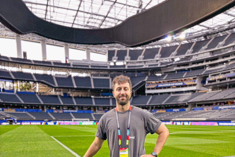 Ein Mann mit Akkreditierung steht lächelnd am Spielfeldrand eines leeren Fussballstadions.