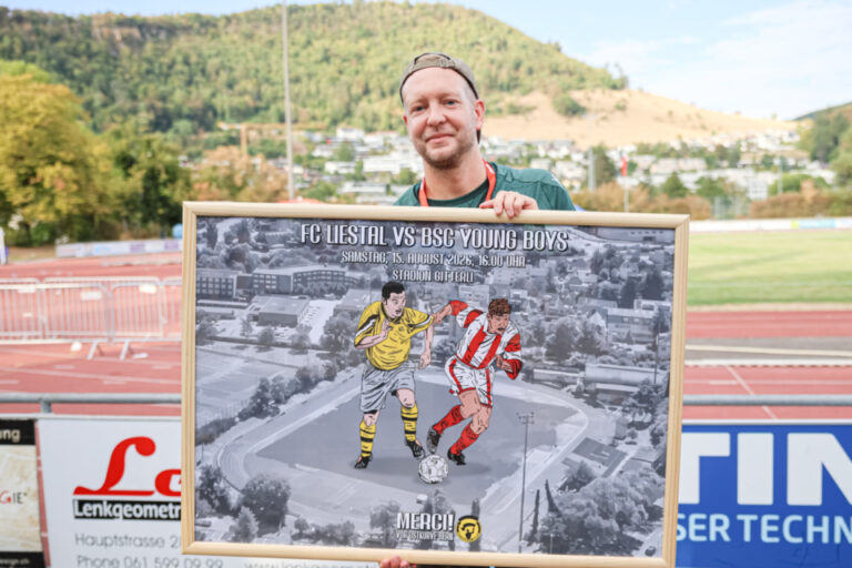 Ein Mann mit Cappy hält ein gerahmtes Bild mit zwei Fussballspielern vor einem Fussballstadion.