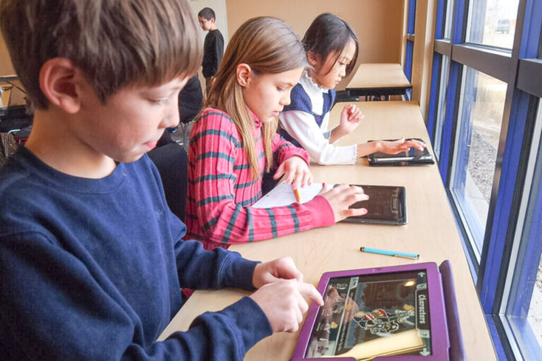 Drei Kinder sitzen nebeneinander an einem Tisch und bedienen Tablets mit den Fingern.
