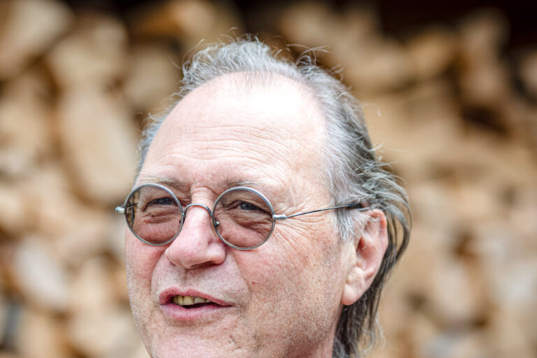 Ein älterer Mann mit runder Brille und grauem Haar lächelt vor einem Holzstapel im Hintergrund.