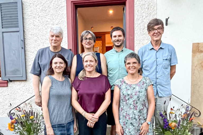 Sieben Personen stehen und knien für ein Gruppenfoto vor einem Hauseingang mit Blumensträussen.