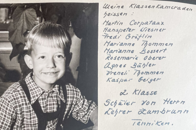 Ein lächelnder kleiner Junge im Karohemd mit Hosenträgern sitzt vor einem aufgeschlagenen Bilderbuch, daneben handgeschriebene Klassenliste.