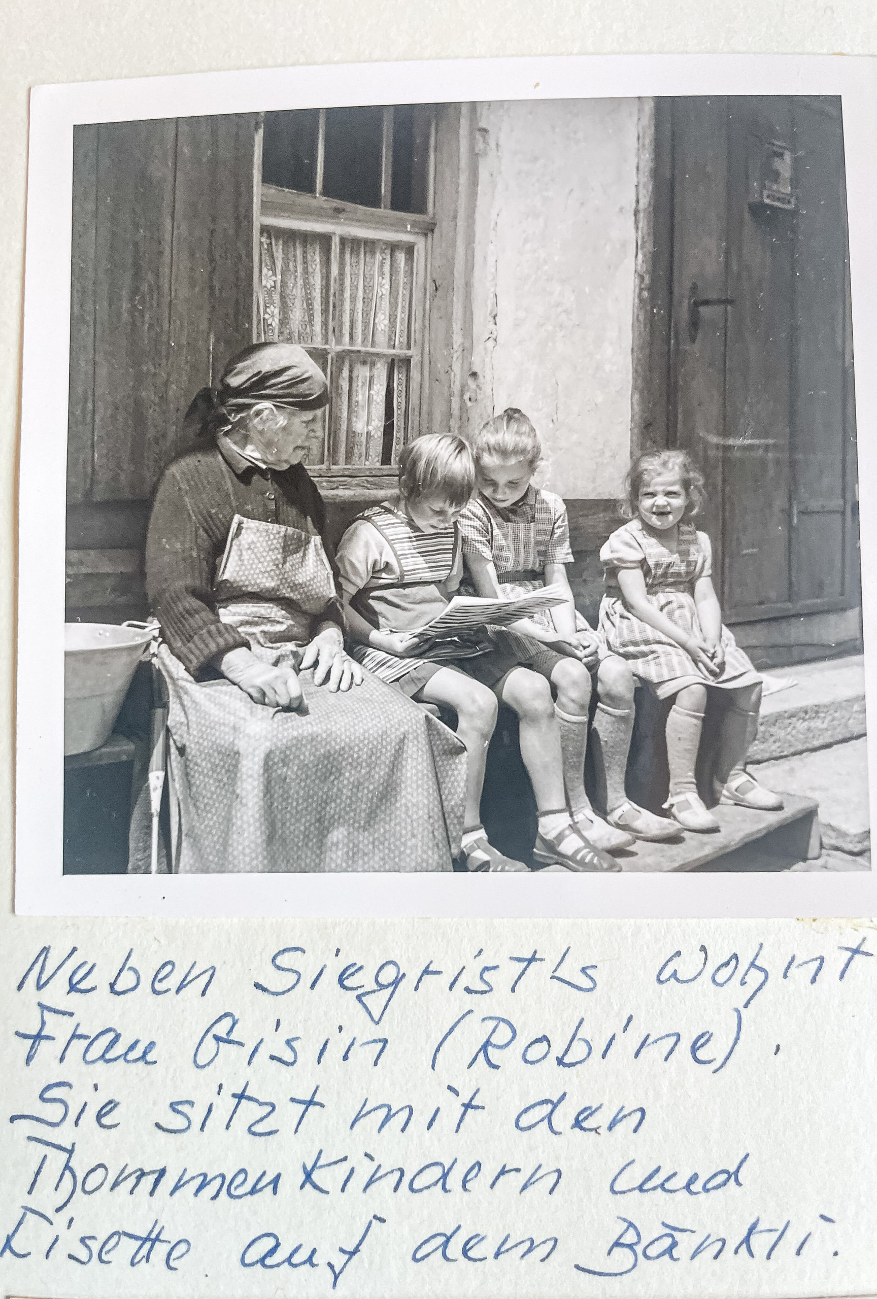 Eine ältere Frau sitzt mit drei Kindern auf einer Bank vor einem Haus und schaut in ein Buch.