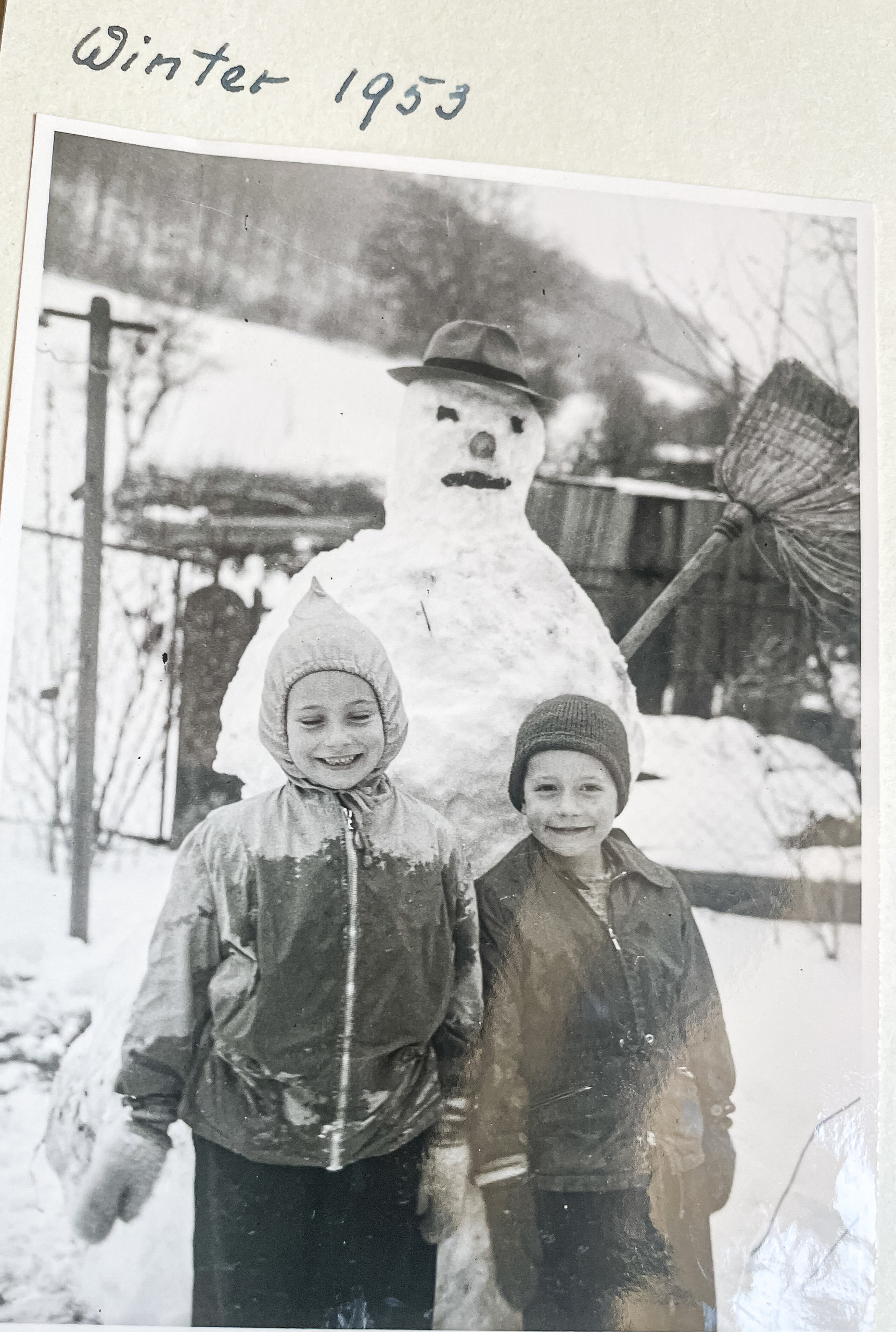 Zwei lächelnde Kinder in Winterkleidung stehen neben einem Schneemann mit Hut im verschneiten Garten.