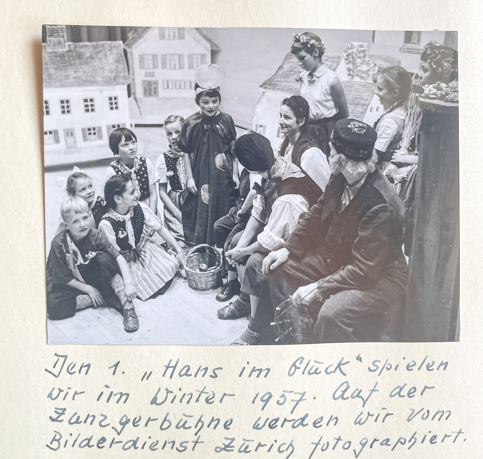 Kinder und Erwachsene in historischen Trachten sitzen und stehen vor einer Kulisse mit Miniaturhäusern.