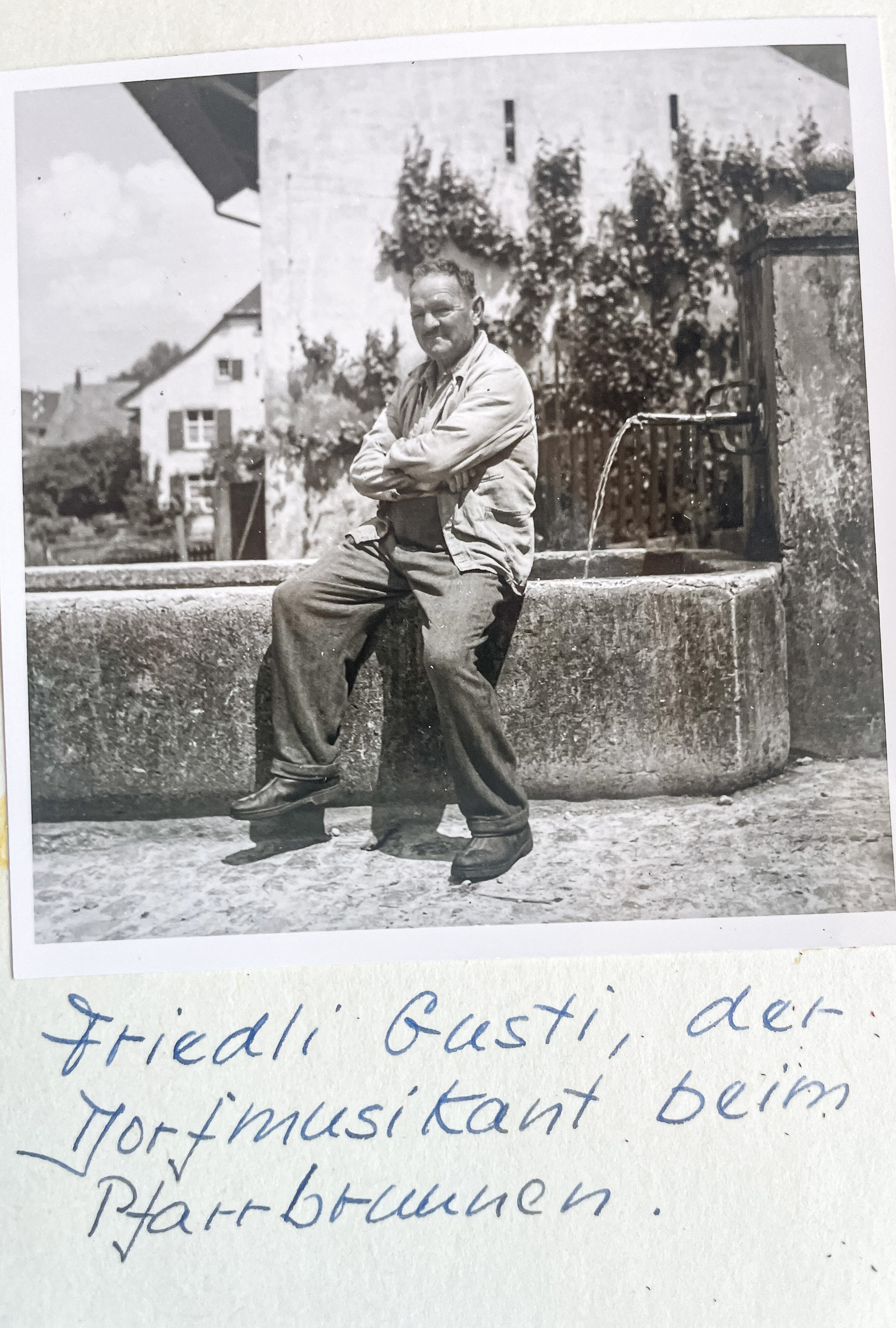 Ein Mann mit verschränkten Armen sitzt lächelnd auf dem Rand eines steinernen Dorfbrunnens.