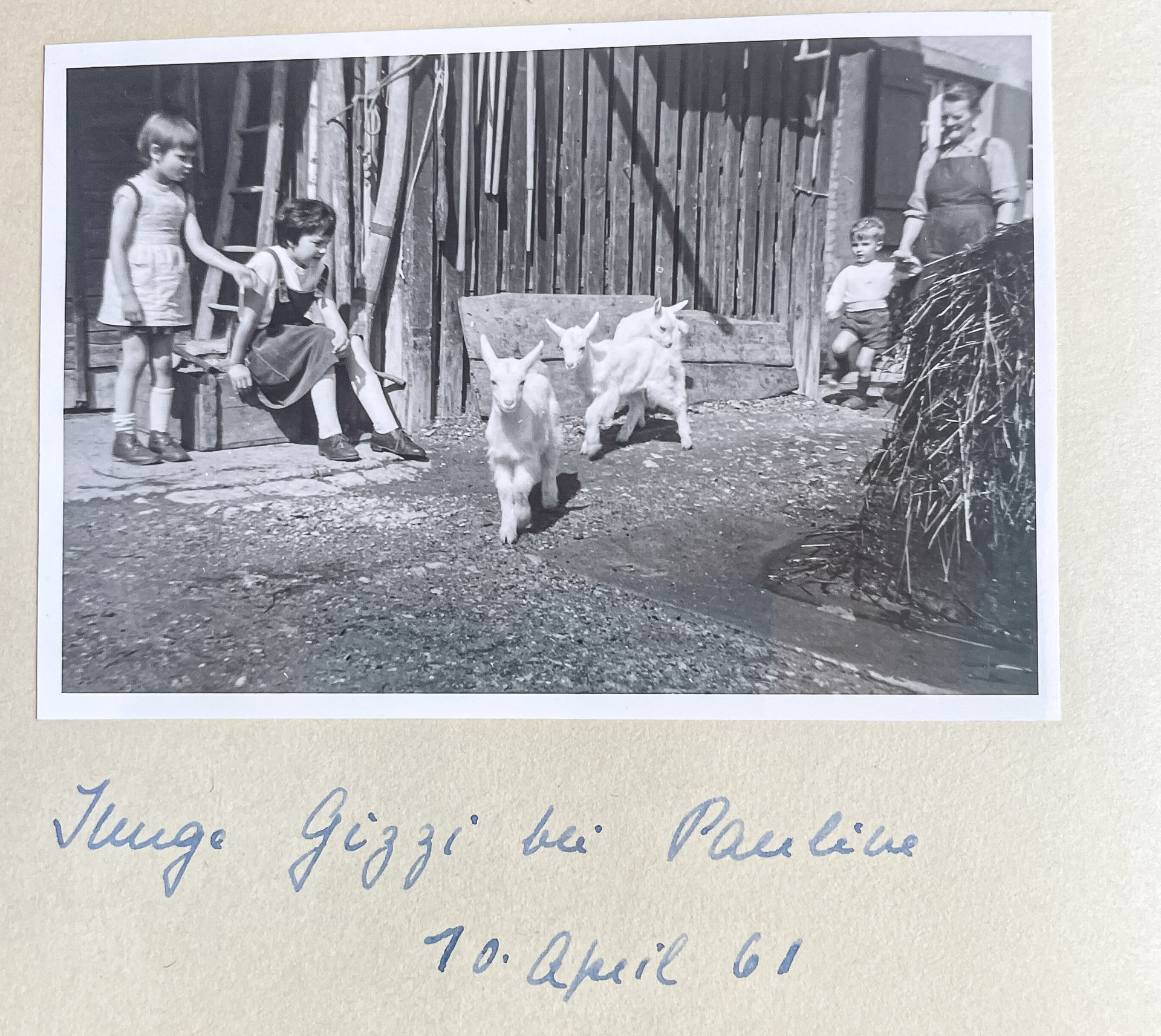 Altes Schwarzweißfoto: Kinder spielen mit zwei Ziegen im Hof vor einer Holzscheune, eine Frau schaut zu.