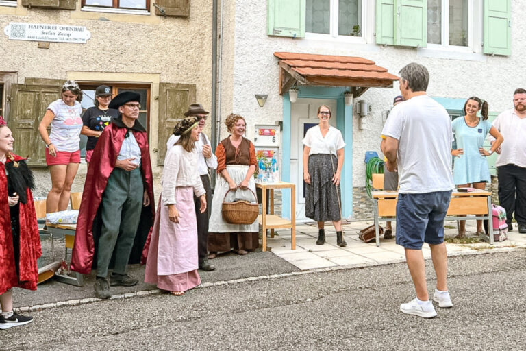 Menschen in historischen Kostümen stehen vor Häusern in einer Dorfstrasse, ein Mann filmt sie.