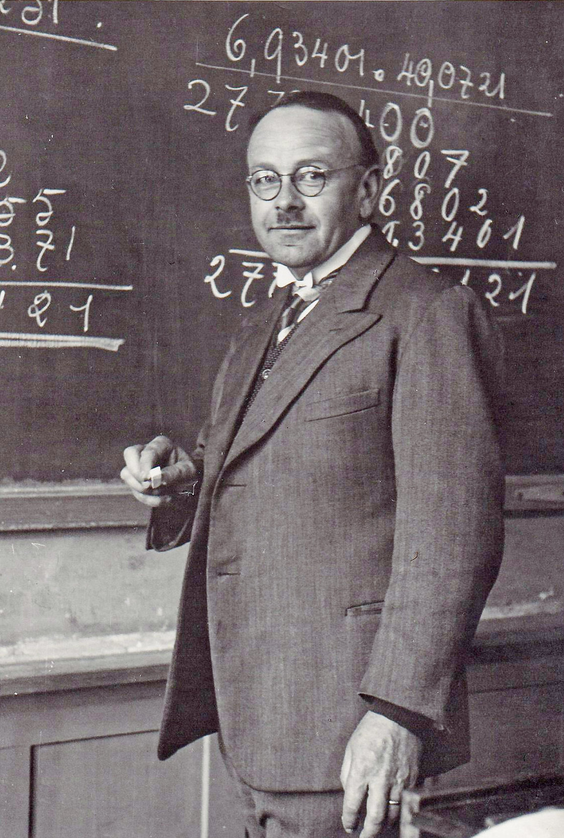 Ein Mann mit Brille und Anzug steht vor einer Tafel mit Rechenaufgaben und hält Kreide in der Hand.