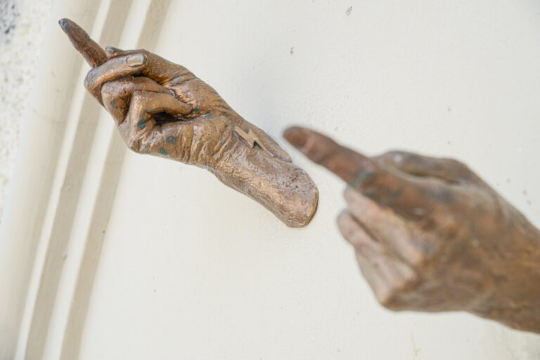 Zwei bronzene Hände strecken sich mit ausgestrecktem Zeigefinger einander entgegen, ohne sich zu berühren.
