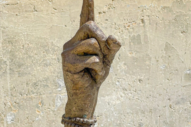 Eine bronzene Skulptur einer erhobenen Hand mit Armreif, auf deren Zeigefinger eine kleine Figur balanciert.