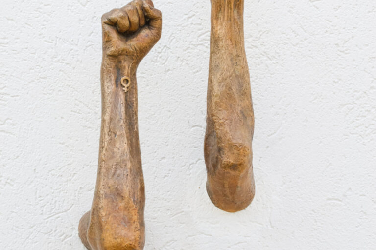 Zwei bronzene, erhobene Fäuste ragen aus einer weissen Wand hervor.